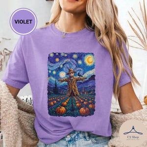 Puede incluir: Una camiseta violeta con un estampado gráfico de un espantapájaros en un campo de calabazas bajo un cielo estrellado y arremolinado. La obra incorpora tonos vibrantes de azul, amarillo y naranja, con un cuervo posado en el hombro del espantapájaros.
