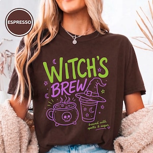 Può includere: Una t-shirt a maniche corte marrone scuro con la scritta Witch's Brew in verde e viola. La grafica include un calderone viola con un teschio, un cappello da strega viola su una tazza e la scritta brewed with spells & sugar in piccole lettere verdi.