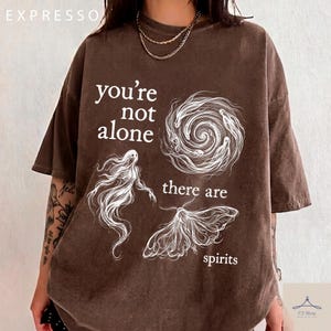 Könnte beinhalten: Ein braunes Kurzarm-T-Shirt mit weißen Linienzeichnungen eines wirbelnden Vortex, eines Geistes und einer Motte. Der Text lautet, you're not alone there are spirits. Das T-Shirt hat einen Rundhalsausschnitt und eine verwaschene Vintage-Optik.