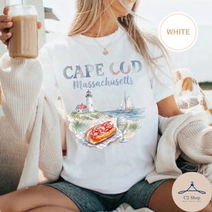 Puede incluir: Una camiseta blanca con un gráfico estilo acuarela y el texto "CAPE COD Massachusetts". El diseño incluye un faro, un velero en el agua, un sándwich de langosta sobre una servilleta a cuadros y una copa de vino blanco en la playa.