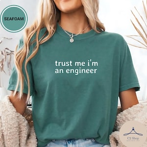 Puede incluir: Camiseta verde mar con el texto blanco "trust me i'm an engineer". La camiseta tiene cuello redondo y mangas cortas. La modelo lleva un collar plateado. La camiseta es de material suave.