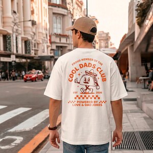 Puede incluir: Una camiseta blanca con un gráfico naranja en la espalda. El gráfico presenta un taco de dibujos animados con un sombrero y el texto "COOL DADS CLUB". El texto adicional incluye "EST 2026" y "POWERED BY LOVE & DAD JOKES".