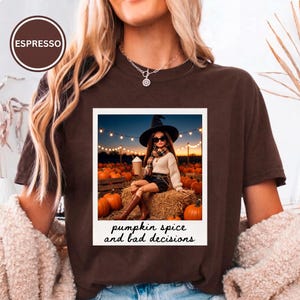 Puede incluir: Una camiseta de manga corta de color marrón oscuro con un estampado gráfico rectangular de una muñeca con sombrero de bruja, gafas de sol y suéter, sentada sobre balas de heno en un campo de calabazas. El texto dice: "pumpkin spice and bad decisions."