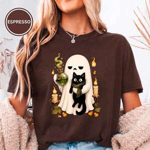 Op de afbeelding: Een donkerbruin t-shirt met een afbeelding van een witte geest die een zwarte kat met een groene sjaal vasthoudt. De geest houdt een groen toverdrankflesje vast, omringd door brandende kaarsen, herfstbladeren en snoepjes. De tekst ESPRESSO staat in een cirkel.