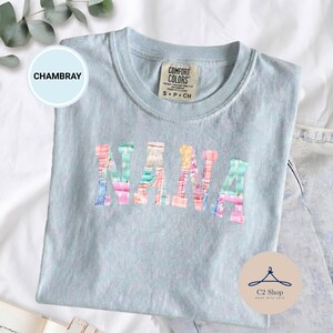 Puede incluir: Una camiseta Comfort Colors azul claro con la palabra "NANA" en letras de patchwork coloridas. La camiseta tiene cuello redondo y mangas cortas. La etiqueta dice "S P CH". La camiseta está sobre una superficie blanca.