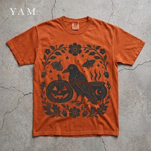 May include: A burnt orange short-sleeve t-shirt featuring a dark charcoal graphic print of a crow, a jack-o'-lantern, a steaming cauldron, moths, and floral elements arranged in a circular wreath pattern on the front of the garment.