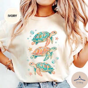 Puede incluir: Una camiseta de color marfil con un estampado gráfico de tres tortugas marinas nadando verticalmente, rodeadas de pequeñas estrellas de mar y burbujas. Las tortugas están ilustradas en tonos turquesa, naranja y amarillo. El texto IVORY aparece en un círculo.