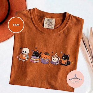 Puede incluir: Una camiseta de color naranja quemado con un estampado gráfico que muestra un esqueleto, dos gatos negros, una calabaza y una taza de café, todos leyendo libros. La etiqueta interior del cuello dice Comfort Colors S P CH.