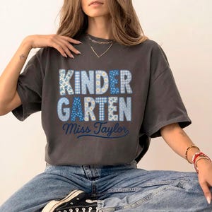 Puede incluir: Una camiseta de manga corta gris carbón con la palabra KINDERGARTEN en letras grandes y gruesas rellenas de patrones florales y de cuadros azules y blancos, con el nombre Miss Taylor escrito en cursiva azul oscuro debajo.
