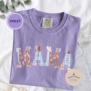 Puede incluir: Una camiseta violeta de manga corta con la palabra MAMA en letras grandes de bloque en el pecho. Las letras tienen un diseño de patchwork con patrones florales y de cuadros en tonos azul, marrón y rosa. Se ve una etiqueta de la marca Comfort Colors.