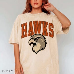 May include: An ivory-coloured, acid-washed short-sleeve t-shirt featuring the word HAWKS in large, distressed orange block letters arched above a graphic illustration of a hawk head in shades of brown, black, and cream with a cracked texture.