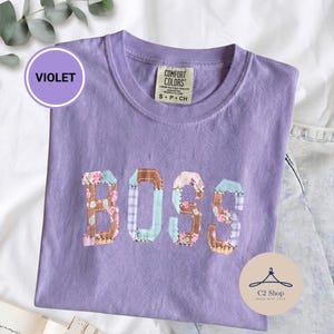 Puede incluir: Una camiseta violeta de manga corta con cuello redondo. En la parte delantera aparece la palabra BOSS en letras grandes de estilo bloque, hechas con un mosaico de telas con estampados florales, de cuadros y de color marrón sólido.