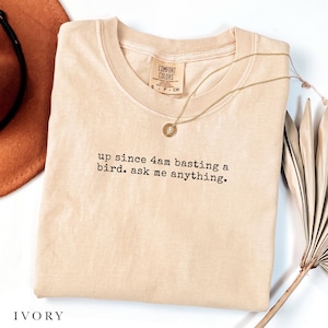 Puede incluir: Una camiseta de color beige claro con cuello redondo y el texto bordado en negro "up since 4am basting a bird. ask me anything." La prenda incluye una etiqueta de la marca Comfort Colors en el cuello y se acompaña de un collar de cadena dorada.