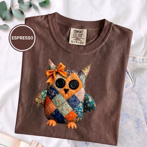 Puede incluir: Una camiseta color espresso con un gráfico de un búho de patchwork que lleva un lazo naranja brillante en la cabeza. El búho está construido con varios patrones de tela florales y geométricos en tonos naranja, azul y verde, con ojos de botón negros.