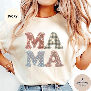 May include: An ivory t-shirt featuring the word MAMA printed in large letters. Each letter has a distinct pattern: the first M is floral, the first A is plaid, the second M is polka dot, and the second A is a different floral print.