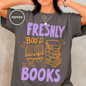 Puede incluir: Una camiseta gris carbón con el texto "FRESHLY BOO'D BOOKS" en morado claro. El gráfico incluye un libro abierto, una pila de libros, gafas naranjas y pequeñas ilustraciones de fantasmas con el texto "best enjoyed after midnight".