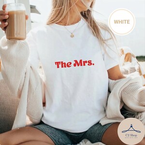 Puede incluir: Camiseta blanca con el texto rojo "The Mrs.". La camiseta es de cuello redondo. La palabra "WHITE" se muestra en un círculo en la esquina superior derecha de la imagen.