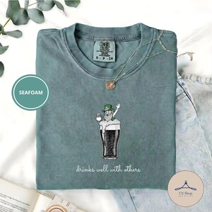 Könnte beinhalten: Seegrünes T-Shirt mit einer Grafik eines Leprechauns in einem Pint Stout-Bier. Der Text "drinks well with others" steht unter der Grafik. Das Shirt hat einen Rundhalsausschnitt und kurze Ärmel. Eine silberne Halskette mit Anhänger befindet sich auf dem Shirt.
