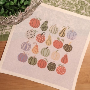 May include: A square needlepoint canvas featuring a grid of seventeen embroidered pumpkins and gourds in various shapes, sizes, and colors including orange, green, yellow, and purple, with some featuring polka dot and checkered patterns.