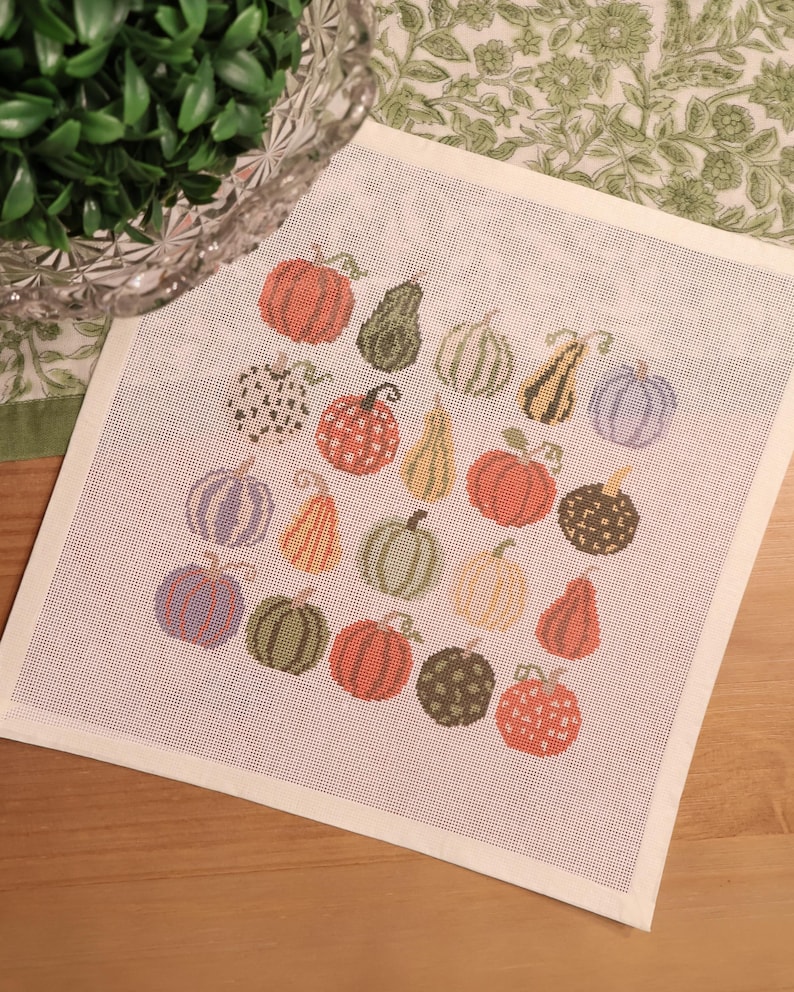 May include: A square needlepoint canvas featuring a grid of seventeen embroidered pumpkins and gourds in various shapes, sizes, and colors including orange, green, yellow, and purple, with some featuring polka dot and checkered patterns.