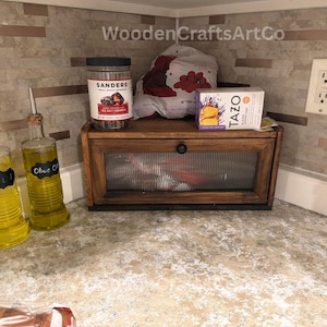 May include: A wooden bread box with a glass front sits on a countertop, next to two olive oil bottles and a jar of sea salt caramels. A box of Tazo tea and a bag of red fruit are also visible.