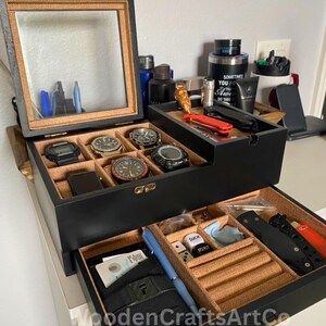 May include: A black wooden watch and accessory organizer with a glass top. The top section holds watches, while the drawer contains a pen, belt, and other items. The interior is lined with a tan fabric.