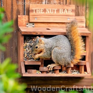 May include: A wooden squirrel feeder with the text "THE NUT BAR" on top. A gray squirrel with a bushy, orange-tipped tail is inside, eating nuts. The feeder is mounted on a wooden fence.