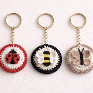 Peut inclure: Trois porte-clés crochetés à la main avec des motifs d'insectes. L'un représente une coccinelle rouge et noire, un autre une abeille noire et blanche, et le dernier un papillon beige. Chaque porte-clés a un anneau et une chaîne dorés.