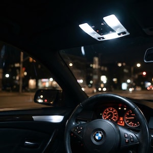 Może przedstawiać: Wnętrze pojazdu marki BMW w nocy, z czarną skórzaną kierownicą z logo BMW, podświetlanymi na pomarańczowo wskaźnikami deski rozdzielczej oraz jasnym białym oświetleniem wnętrza odbijającym się w przedniej szybie i elementach wykończenia.