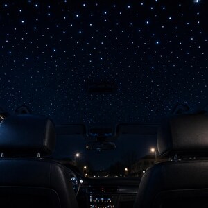 May include: A vehicle interior featuring a black headliner illuminated by numerous small, bright white fiber optic lights that mimic a starry night sky. The cabin includes two black leather headrests and a dark dashboard visible in the background.