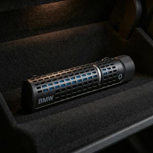 Puede incluir: Un ambientador cilíndrico negro de la marca BMW con una carcasa exterior de patrón de rejilla que deja ver componentes interiores azules. El dispositivo tiene una tapa negra sólida, un anillo plateado y el logotipo de BMW en el lateral.