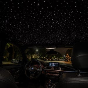 May include: The interior of a vehicle featuring a black headliner illuminated with numerous small, bright white fiber optic lights that mimic a starry night sky. The dashboard, steering wheel, and black leather seats are visible below.