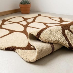 Op de afbeelding: Beige en bruin patroonkleed met een uniek ontwerp. Het kleed heeft een herhalend patroon van bruine vormen tegen een beige achtergrond. Het kleed is gedeeltelijk opgerold, waardoor de onderkant zichtbaar is.