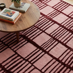 May include: A rectangular area rug featuring a repeating grid pattern of deep burgundy lines set against a soft, muted pink background. The design consists of thick, dark-colored borders framing squares of a lighter, solid pink textile material.