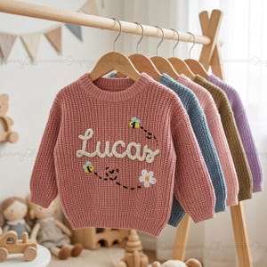 Könnte beinhalten: Eine Reihe von Kinderpullovern in verschiedenen Farben, die auf einem Holzständer hängen. Der vordere Pullover ist rosa und mit dem Namen "Lucas" in Weiß bestickt, zusammen mit Bienen- und Blumenmotiven. Weitere Pullover sind blau, rosa, grün und lila.