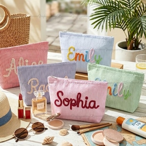 Puede incluir: Varias bolsas de cosméticos personalizadas a rayas en colores como rojo, azul y rosa, con nombres bordados. Las bolsas se exhiben con maquillaje y accesorios, sugiriendo un tema de viaje o belleza.