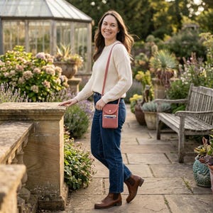May include: A coral-colored crossbody phone bag with a flap closure and a shoulder strap. The bag is worn over a cream-colored sweater and blue jeans. The background features a garden with stone pathways and lush greenery.