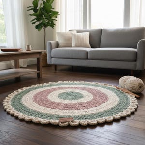 May include: A circular, hand-crocheted rug with a scalloped border. The rug displays concentric rings in green, pink, and cream. A ball of yarn and a crochet hook are beside the rug. The rug is placed on a dark wooden floor.