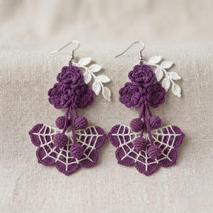 May include: A pair of dangle earrings featuring dark purple crocheted flowers and small spheres suspended above a white and purple crocheted spiderweb motif, accented with a white crocheted leaf sprig attached to the silver-toned metal hooks.