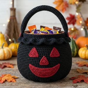 May include: A black crocheted basket shaped like a jack-o'-lantern, featuring a rounded handle, a ruffled top edge, and red crocheted eyes, nose, and a wide smiling mouth. The basket is filled with various small, colorful candy wrappers.