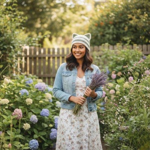 May include: A woman wearing a light blue denim jacket and a floral print dress, holding a bouquet of lavender. She is also wearing a white and green cat ear hat. The background features a garden with a wooden fence and colorful flowers.