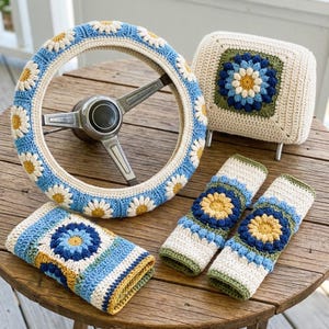 May include: A crocheted car accessory set featuring a steering wheel cover, headrest cover, and seat belt covers. The set is cream-colored with blue, yellow, and green daisy and floral designs. The steering wheel cover is wrapped around a silver steering wheel.