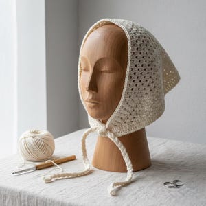 Op de afbeelding: Crèmekleurige gehaakte muts met strikbanden, tentoongesteld op een houten hoofd. Een bol garen, een haaknaald en een kleine schaar liggen naast de muts. De muts heeft een vierkante vorm en een open weefsel.