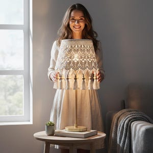 Op de afbeelding: Een beige macram&eacute; tafellamp met een houten voet en een decoratieve lampenkap. De lampenkap heeft ingewikkelde geweven patronen, kwastjes en houten kralen. De lamp staat op een ronde tafel naast een raam.