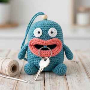 May include: A teal crocheted key holder shaped like a cartoon monster with large pink lips and white eyes. A silver key and key ring are attached. A wooden bead and teal cord are at the top. A ball of yarn and crochet hook are in the background.
