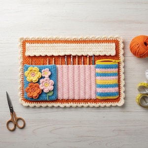 Puede incluir: Un organizador de ganchillos hecho a mano en naranja, crema y azul. El organizador presenta un bolsillo azul con aplicaciones de flores, un soporte para ganchillos rosa y una bolsa con cremallera a rayas. También se ven una madeja de lana naranja y unas tijeras.