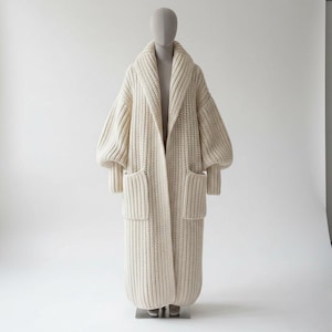 Pode incluir: Um casaco de malha creme oversized com gola de xale e bolsos grandes. O casaco comprido apresenta um design de malha grossa, mangas largas e bainha até ao joelho. O casaco é exibido num manequim sobre um fundo branco.