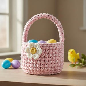 May include: A pink crocheted Easter basket with a white and yellow flower. The basket contains colourful Easter eggs. A yellow chick figurine and greenery are next to the basket. The basket has a handle.