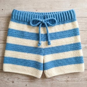 May include: Handmade crochet shorts in a blue and cream striped pattern. The shorts feature a drawstring waist with a bow detail. The crochet work is visible, showcasing the texture and craftsmanship of the garment. The shorts are designed for casual wear.