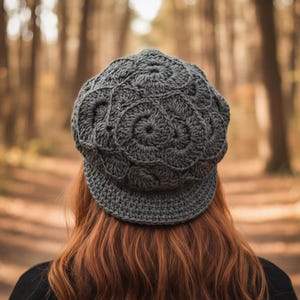 May include: A grey crocheted newsboy cap with a circular design. The cap features a brim and is worn by a person with red hair. The background shows a blurred forest with trees and a path.